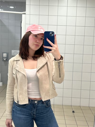 A young woman takes a selfie in a tiled bathroom. She wears a pink baseball cap, a white ribbed tank top, and a tan suede jacket. She also wears blue jeans. The subject holds a dark blue cell phone with red nails. A silver cross hangs around her neck. The bathroom has white tile walls and floor. There are light switches and a hand dryer visible on the wall behind her.