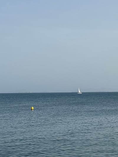 The image depicts a wide, calm expanse of deep blue ocean under a hazy, light blue sky. In the foreground, a bright yellow buoy floats on the water's surface. Further out towards the horizon, a solitary white sailboat with its sail raised is visible, moving slowly across the sea. A faint, indistinct shoreline with some structures is visible in the far distance, suggesting the coast of Urbanización Cabo Roig, Spain. The overall impression is one of tranquility and open space.