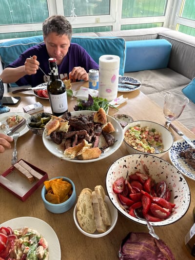 A group of people are gathered around a wooden table, enjoying a meal. The man in the foreground, wearing a purple t-shirt, is eating from a plate of food. Various dishes are spread across the table, including a large platter of grilled meat, bowls of salad, sliced tomatoes, pickles, bread, and a bowl of tortilla chips. A bottle of wine and a can of Corona Extra are also visible. The scene appears to be set in a sunroom or enclosed patio, with large windows looking out onto greenery. The overall atmosphere is one of a casual, convivial gathering. The image was taken in the city of Ptichnoye, Russia.