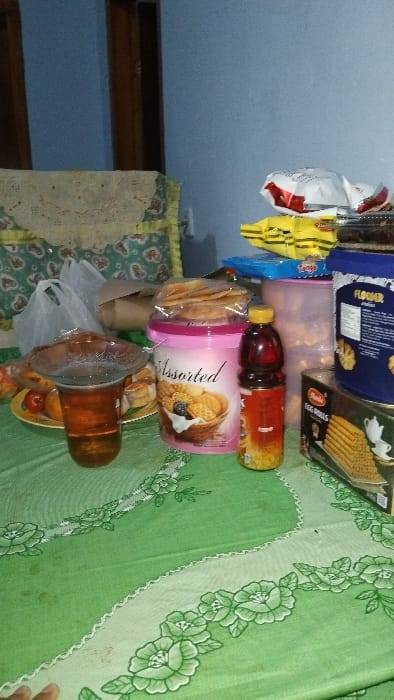 A table with a green tablecloth with a white lace trim and floral pattern is set with a glass of brown liquid, a pink plastic container of assorted cookies, a glass jar of cookies, a red and white plastic container with snacks, a clear glass jar of cookies, and a brown glass bottle of liquid. There are three plastic bags in the background, a partially obscured brown object, and a chair with a white and beige patterned cushion. The location is in Panjalu, Indonesia, at coordinates (latitude: -7.142, longitude: 108.277). The scene is indoors and appears to be a living room or dining room.