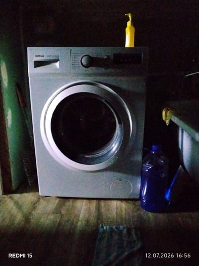 A front-load washing machine is centrally positioned in the frame. It is a light gray appliance with a circular glass door and a control panel on the top left. Above the washing machine, a yellow liquid soap dispenser is visible. To the right of the washing machine, a large blue plastic bottle is placed on a slatted surface, possibly a laundry basket or shelf. The floor appears to be made of wooden planks. The background is dark and indistinct. The lower left corner of the image displays "REDMI 15" and the lower right corner shows "12.07.2026 16:56", indicating the device and date/time the photo was taken. The location is not specified in the image.