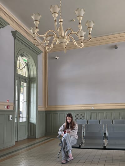 A young woman sits in a waiting area in Przemyśl, Poland. She is wearing a light-colored hooded sweatshirt, grey pants, and pink shoes. She is seated on a metal chair with a chrome frame. The waiting area has metal seating aligned against a wall. The wall is painted a light green color. In the background, there is a large window and door structure. A chandelier hangs from the ceiling. The floor is tiled. A security camera is mounted on the ceiling.