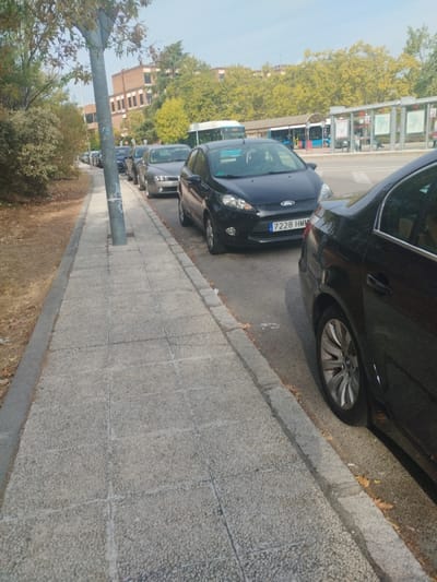 The image captures an urban street scene in Pozuelo de Alarcón, Spain, featuring a concrete sidewalk, a line of parked cars, and a road with a bus stop. In the foreground, a textured, light grey concrete paver sidewalk extends from the bottom left to the upper center. To the left of the sidewalk is a narrow strip of dry earth with sparse vegetation and fallen leaves. A grey utility pole is positioned on the sidewalk in the middle ground.
To the right of the sidewalk, several vehicles are parked parallel to the curb. The rear passenger side of a dark-colored sedan, equipped with a multi-spoke silver alloy wheel, is prominent in the immediate right foreground. Ahead of this vehicle, a black Ford Fiesta hatchback with Spanish license plate 7228 HMM is clearly visible. Further down the street, a silver-grey sedan and additional vehicles are parked in a continuous line.
Across the asphalt road, a public bus stop with multiple shelters and information panels is present. A blue and white bus is partially visible at the stop. Behind the bus stop and road, a dense row of trees, displaying both green and some yellowing foliage, creates a natural barrier. In the distant background, a large, multi-story building constructed of light-colored brick with prominent horizontal window bands is visible. The sky above is clear and light blue. No individuals are discernible, and no active interactions are depicted within the scene.