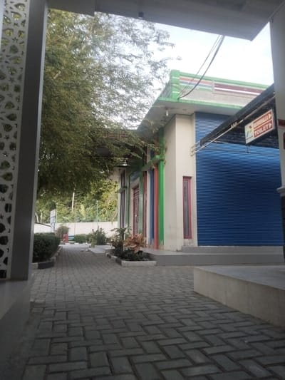 The image depicts an outdoor walkway in Glee Gogo, Indonesia, leading towards a building with a blue roller shutter door. To the right of the building, a sign indicates "MUSHOLLA" and "TOILET" with an arrow. The walkway is paved with interlocking bricks. Lush green trees and foliage are visible on the left side of the walkway and in the background. The building itself appears to be a single-story structure with light-colored walls and colorful vertical trim. The sky is overcast. No people are present in the scene.