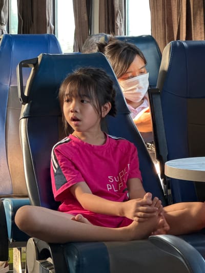 The image shows two young girls sitting in a bus, which is likely in motion given the blurriness of the background. The bus appears to have blue seats, and the girls are seated one behind the other. The girl in the foreground, wearing a pink t-shirt with white stripes on the sleeves, is looking towards the camera with a surprised expression. Her arms are clasped together. The girl in the background is wearing a white face mask and her head is turned away from the camera, resting against the seat. The lighting suggests it is daytime, possibly late afternoon due to the warm tones. The curtains on the windows are a textured brown. The scene is set in Hòa Đa, Vietnam. There is no visible text in the image.