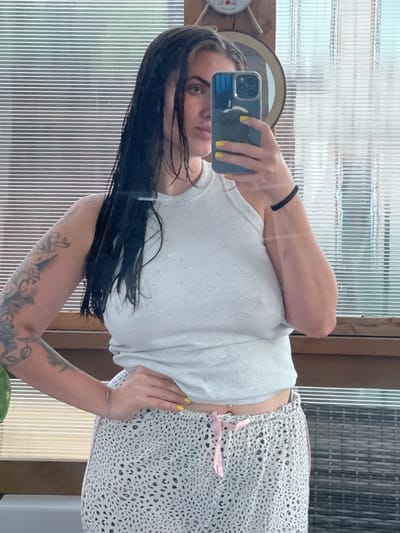 A woman is taking a selfie in a mirror. She has long, dark hair and is wearing a grey tank top and white patterned shorts with pink ties. She has yellow nail polish on her fingers and a nose ring. Her arm is resting on her stomach, and she has tattoos on her arm. The background appears to be a room with ribbed glass windows and wooden accents. A thermometer is visible on the left side of the frame. The lighting suggests it is daytime, and the overall atmosphere is casual and relaxed. The image is rotated 90 degrees counter-clockwise.