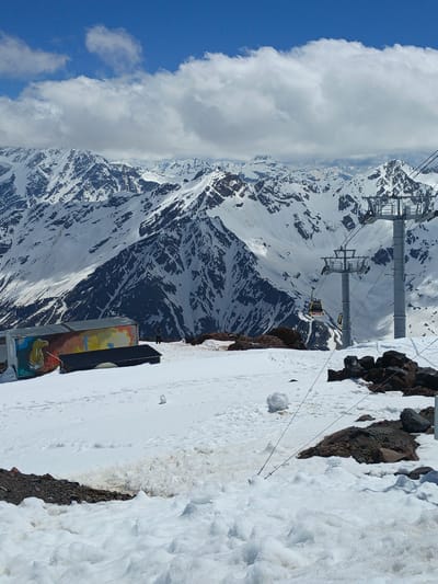 The image captures a scenic view of the snowy mountains surrounding Terskol, Russia. In the foreground, a ski lift with yellow cabins ascends towards the snow-capped peaks. A building with colorful murals is visible to the left, and a person stands near it. The terrain is covered in snow, with rocks and debris scattered across the ground. The sky is a vibrant blue, with scattered clouds adding depth to the scene. The image appears to be taken on a clear day, with ample sunlight illuminating the landscape. The overall impression is one of a breathtaking mountain vista, likely a popular spot for skiing and other winter sports.