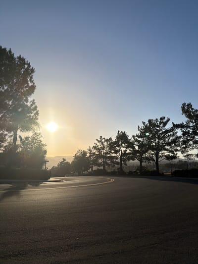 The image captures a view of a curved asphalt road with yellow lane markings in Aliso Viejo, California, USA. The sun is setting on the left side of the frame, casting a warm glow and lens flare across the scene. Silhouetted trees, including pine trees, line the right side of the road and form a dark border against the clear, pale blue sky. Distant hills are visible in the background, softened by the haze of sunset. The road surface appears to be in good condition, with some reflections of light on its surface. There are no people or vehicles visible in the image. The overall atmosphere is serene and natural, typical of a suburban or semi-rural setting.