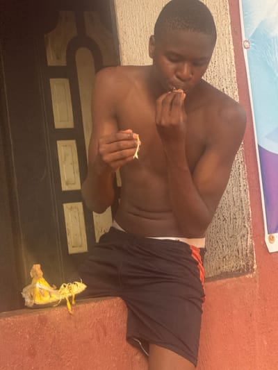 A young, dark-skinned man is depicted in a reclined or seated position, consuming a light-colored food item with his hands. He appears shirtless, wearing dark shorts with a visible orange stripe along the side and a white waistband. He holds pieces of the food in both hands, with one hand bringing a morsel to his mouth.

The man is positioned on a reddish-brown, textured surface, possibly a concrete step or ledge. The ground directly in front of him features a dark, possibly tiled, surface with lighter rectangular patterns. To his lower right, near his feet, lies discarded refuse, appearing to be a yellow or light brown fruit peel or wrapper.

In the upper background, a wall or structure with a similar reddish-brown base is visible. Above this base, a banner or poster with sections of light blue, white, and purple/dark blue is displayed. No discernible text is visible on the banner or elsewhere in the image.

The scene suggests an outdoor or semi-outdoor setting, with bright lighting indicating it is daytime. The man's expression is neutral and focused on eating, conveying a sense of calm. The location context for the image is Aguata, Nigeria.