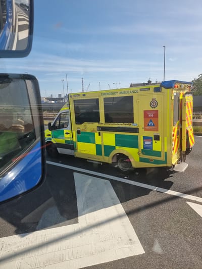 An ambulance, marked "EMERGENCY AMBULANCE" and "South Central Ambulance Service NHS", is stopped on a road with white lines, possibly at a junction or intersection, under a clear blue sky with some light clouds. The ambulance is yellow and green with reflective markings. A blue vehicle's side mirror and part of its body are reflected in the foreground, suggesting the photo was taken from another vehicle. In the background, urban infrastructure, including buildings, streetlights, and cranes, is visible. The road surface appears to be asphalt with painted white markings. The scene is static, with no apparent activity beyond the parked ambulance.
