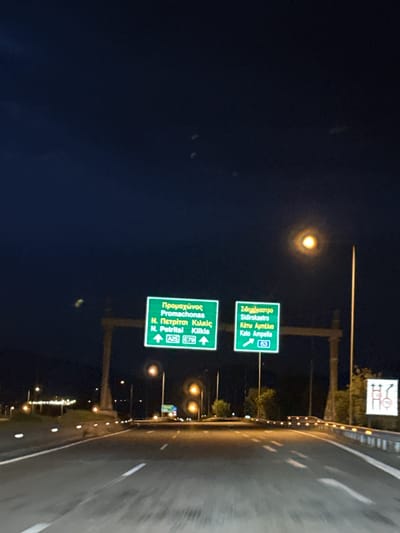 The image shows a road at night in or near Sidirokastro, Greece. Two green highway signs are visible above the road. The top sign indicates directions for Sidirokastro and Kato Ampelia (Route 63). The bottom sign indicates directions for Promachonas, N. Petritsi, and Kilkis (A25 and E79). The road has reflective lane markers. Streetlights are visible along the side of the road.