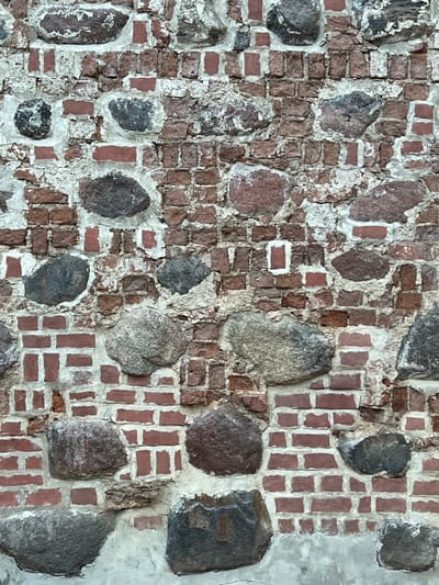 A detailed, forensic close-up depicts an aged, vertical wall surface located in Мірскі сельскі Савет, Belarus. The wall is constructed from an irregular mixture of large, rounded to ovate natural stones, ranging in color from dark grey to various shades of brown and beige, interspersed with numerous smaller, reddish-brown bricks. The stones are set within a substantial matrix of light-colored mortar, which exhibits signs of significant wear, erosion, and discoloration, appearing rough, cracked, and partially degraded in many areas. The bricks, often appearing as fragmented or incomplete units, are laid in irregular courses or as infill between the larger stones, contributing to the wall's uneven texture. Portions of the mortar have crumbled, exposing deeper recesses or the edges of the construction materials. The overall scene shows a robust, historical construction with visible material degradation and weathering. No individuals, discernible actions, or external objects are present within the frame, focusing solely on the structural elements and their condition.