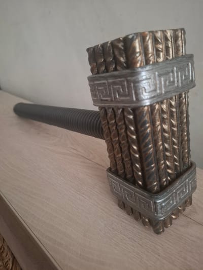 This media file shows a close-up, angled shot of a prop hammer, likely made of metal rebar and painted to appear aged. The hammer head is composed of many vertical, twisted metal rods bound together by two decorative bands. These bands have a Greek key or meander pattern embossed on them, suggesting a historical or fantasy theme. The handle of the hammer is a ribbed, dark gray shaft, possibly made of rubber or a similar flexible material. The hammer is resting on a light-colored wooden surface, possibly a table or shelf, with a plain, light-colored wall in the background. The lighting appears to be artificial, casting soft shadows and highlighting the textures of the metal and wood. There is no discernible activity taking place; the image is a still representation of the object. The overall impression is one of craftsmanship and design, with the hammer appearing to be a decorative item or costume prop. There is no visible text in the image. The location context is Bălți, Moldova.