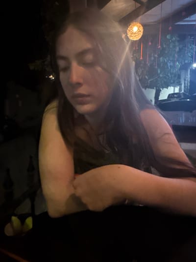 The image depicts a young female, estimated to be in her late teens or early twenties, seated at a dark table in what appears to be an outdoor or semi-enclosed restaurant or bar environment at night. The scene is illuminated by warm, artificial light sources, resulting in areas of deep shadow and bright highlights. The female, positioned center-frame, has long, dark hair that falls over her shoulders. She is wearing a light-colored top, possibly olive green. Her forearms are resting on the dark table surface, with her left hand lightly grasping her right wrist. Her eyes are closed or heavily downcast, and her facial expression is neutral, suggesting repose or introspection.

In the background, overhead, a spherical light fixture made of woven material hangs from a wooden beam. Several red, string-like decorations also hang from above, visible behind the subject. To the left, partially obscured by shadow, two bottles are visible on a dark shelf, and a small cluster of green objects, possibly limes, are illuminated on the table surface. Further back and to the right, green foliage from a tree is visible, and a portion of a dark-colored vehicle, likely a car, is discernible in the far background, indicating proximity to a street or parking area. The overall setting suggests a casual, evening social establishment in the city of Santiago, Mexico.