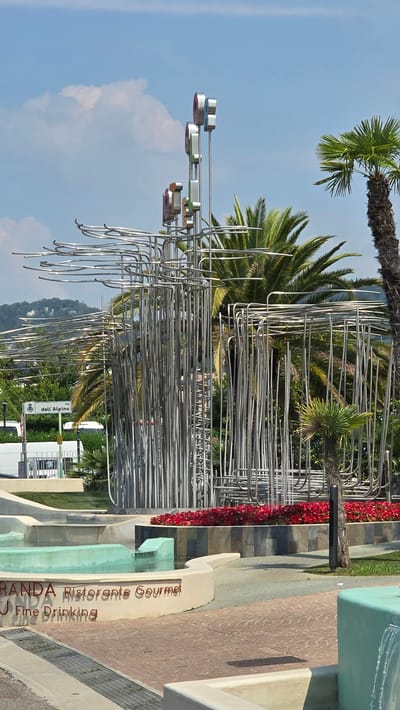 This is a photograph of a public art installation in Bardolino, Italy. The installation features a complex structure made of interconnected metal tubes, with several circular elements at the top that appear to be made of chrome and colored glass or plastic. The sculpture is situated in front of a backdrop of palm trees and lush greenery, with a hillside visible in the distance. In the foreground, there is a modern fountain with turquoise water, surrounded by curved concrete structures. Text is visible on one of the concrete structures, reading "GRANDA Ristorante Gourmet" and "Fine Drinking". A street sign indicating "via dell'Alpino" is also present, suggesting the installation is located near that street. The sky is blue with some scattered clouds. The scene appears to be a public space, likely a plaza or park, designed for leisure and dining, given the presence of the restaurant signage and fountain. There are no people visible in the image.
