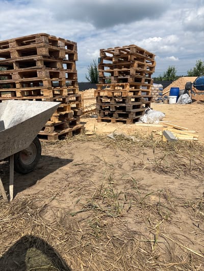 The image depicts a construction site in Opatów, Poland. In the foreground, a concrete wheelbarrow is positioned on sandy ground covered with dry grass and straw. Behind the wheelbarrow, two large stacks of wooden pallets are visible. To the right of the pallets, there are several bags of construction material, a blue barrel, and a concrete mixer. The sky above is partly cloudy with dramatic cumulus clouds. The overall scene suggests an active construction or renovation project.