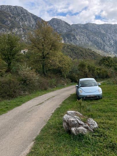 The image depicts a rural mountainous landscape in Đenjaši Česminovo, Montenegro. A light blue, second-generation Volkswagen Beetle, bearing a Russian Federation license plate "X458OT 142," is parked on a grassy verge to the right of a narrow, unpaved or lightly paved road. The road, which shows minor cracking, curves from the lower left towards the upper left-center of the frame. In the immediate foreground on the right, a large, light-colored, irregularly shaped rock rests in the grass. The midground features dense green and autumnal foliage, including several trees with yellowish-green leaves. Faintly visible amidst the trees on the left side are small white buildings with red roofs. The background is dominated by rugged, grey mountains, partially covered in sparse vegetation, beneath a partially cloudy sky. No individuals are present, and no actions or interactions are depicted.