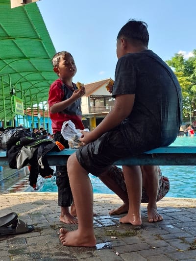 The image depicts two boys, one older and one younger, sitting on a bench near a pool. The older boy is wearing a black shirt, black shorts, and is barefoot. The younger boy is wearing a red shirt, blue shorts, and has bare feet. Both are wearing black sandals, the younger boy’s on his feet and the older boy’s on the bench next to him. The younger boy is holding a piece of food in his right hand. Both boys are looking at each other. The bench is blue, and the pool is blue and clear. The area surrounding the pool and bench is paved with gray bricks, with some green grass growing in between. The image was taken in Kecamatan Tulung, Indonesia, at the coordinates (latitude: -7.592, longitude: 110.649). There is a green awning above the bench, and a building is in the background.
