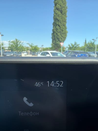 The image shows a close-up of a car's infotainment screen displaying the time as 14:52 and the temperature as 46°C. Below the time and temperature, there is a phone icon and the word "Телефон" (Telephone) in Cyrillic script. The screen appears to be dirty with several smudges and scratches. In the background, out of focus, are cars parked in what appears to be a parking lot, trees, streetlights, and a bright blue sky. A tall, slender cypress tree is prominent in the background to the left of center, with a triangular yield sign visible at its base. The scene suggests a very hot day, possibly in Mojanovići, Montenegro, given the context provided.