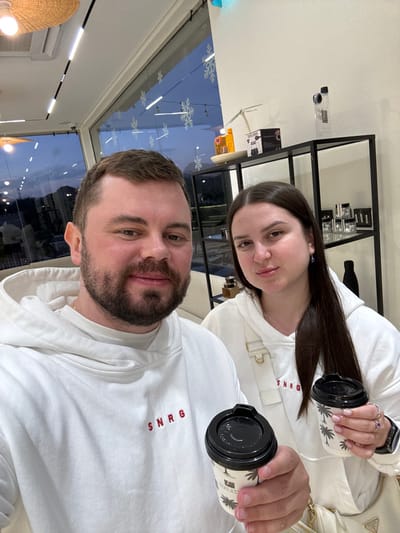 Two individuals, an adult male and an adult female, pose for a photograph indoors at twilight within a cafe or lounge in Hatta, United Arab Emirates. The male, with dark hair and a beard, wears a white hooded sweatshirt embroidered with "SNRG" in red text. He holds a white takeaway coffee cup with a black lid and a palm tree design in his right hand. The female, with long dark hair, wears a matching white hooded sweatshirt with "SNRG" and a light-colored shoulder bag, holding an identical coffee cup in her left hand. Both cups bear "STAR CAFE" branding. The interior features white walls, linear ceiling lighting, a woven light fixture, and large windows adorned with snowflake decals. Through the windows, an outdoor area with distant buildings and lights is visible against a twilight sky. A black metal shelving unit displaying various products, including bottles and boxes, is positioned on the right.