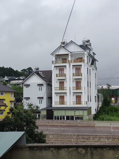 The image captures a view of a modern white hotel building with multiple balconies, located in the city of Lang Biang - Đà Lạt, Vietnam. Adjacent to the hotel is a light gray residential building with a dark roof and several windows. A bright yellow building is visible further to the left. The scene is set against a backdrop of lush green hills and trees, with a slightly overcast sky. In the foreground, there is a rooftop with corrugated metal sheeting and a concrete wall. The overall impression is that of a densely built urban or semi-urban area within a hilly landscape.