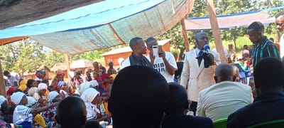 A gathering of people is occurring outdoors in Kalemie, Democratic Republic of the Congo, under a makeshift awning constructed from blue tarps. A man in a light-colored suit is speaking into a microphone. To his left are two younger men, one holding up a mobile phone as if recording or taking a picture. A large crowd of people, primarily women with headscarves, are seated in front of the speaker. In the background are trees and buildings.