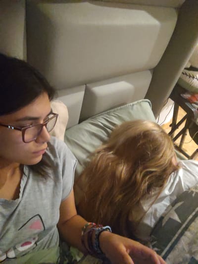 The image depicts an indoor scene, likely a bedroom, with a person wearing glasses and a gray t-shirt with a cat graphic. Beside them, another person with long, reddish-blonde hair is lying down, with their face obscured by their hair. Both individuals appear to be resting on a bed with pillows. The background shows a tufted headboard and a glimpse of a table with items on it. There is no visual information in the image that indicates a specific location in Chorrillos, Peru.