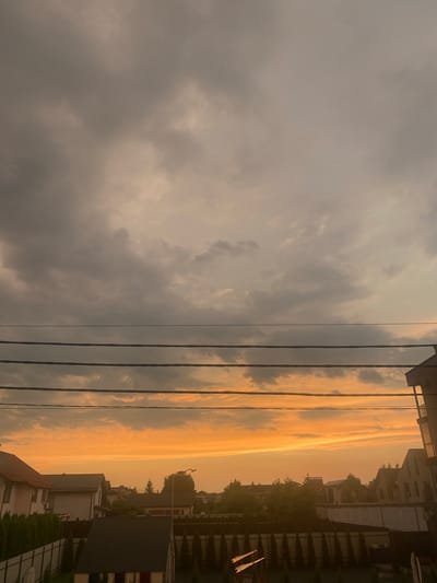 The image depicts a suburban street scene in Novi Petrivtsi, Ukraine, during a sunset. The sky is filled with dramatic clouds, ranging in color from dark gray at the top to a vibrant orange and yellow horizon. Three thick black wires stretch horizontally across the frame, obscuring parts of the sky. Below the wires, a collection of houses with pitched roofs, fences, and lush green trees are visible. The architecture suggests a residential area with varying roof styles and building materials. The lighting indicates the late hour of the day, casting a warm glow over the scene. There are no people or vehicles present in the image.