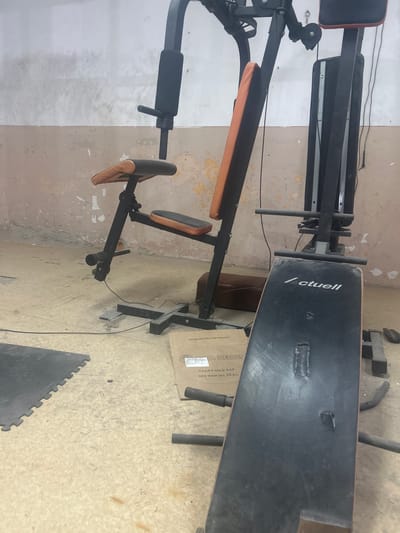 The image shows a home gym setup in an indoor setting. The gym equipment includes a weight bench and a multi-gym machine, both appearing slightly worn. The bench has the word "Actuell" printed on it. The equipment is situated on a floor covered with what looks like interlocking foam mats. A cardboard box with text "A HELLAS" and "VEGETABLE FAT NET WEIGHT 25 KG" is placed next to the bench. The walls are roughly finished with visible signs of wear and old paint. The photo appears to be taken indoors, possibly in a basement or garage. Based on the user's prompt, this scene is presumed to be in Kalchevo, Bulgaria.