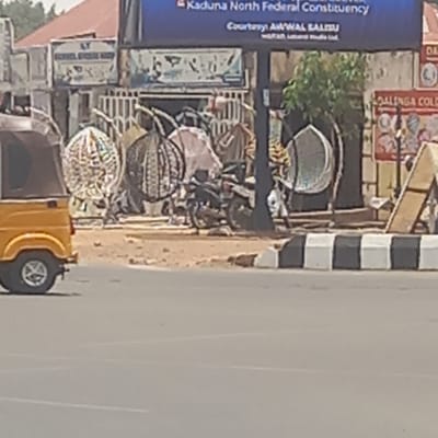 The image depicts an urban street scene in Kabala, Nigeria. A yellow tricycle is parked on the right side of the frame. In the background, there are people, buildings, commercial signs, and merchandise displayed outside shops. Woven baskets are visible amidst other objects. The environment suggests a bustling marketplace or commercial area along a roadside.