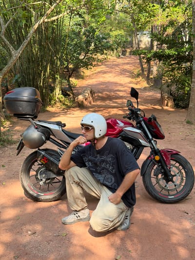 The image shows a man in Thủy Xuân Ward, Vietnam, posed with a red Honda motorcycle. The man wears a white helmet, sunglasses, a dark t-shirt, and khaki pants, and gray shoes. He is leaning on the motorcycle, which is parked on a dirt road or path. The motorcycle has a black storage box mounted on the back. The environment includes trees, bamboo, and a dirt path. The overall setting appears to be a wooded or semi-rural area.