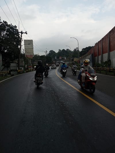 The image depicts a two-lane asphalt road in Dawathang_Dorjibi_Kashingtsawa, Bhutan, under an overcast sky, suggesting recent rain as the road surface is visibly wet. A solid yellow line divides the road longitudinally.

In the foreground and midground, a total of at least nine individuals on motorcycles or scooters are visible. From the low-angle perspective, a dark-clad rider on a dark motorcycle with a visible license plate is positioned in the left lane, moving away from the viewer. Behind this motorcycle, another rider on a similar vehicle follows. In the opposing lane, closer to the viewer, a person on a red scooter wears a light-colored top, light trousers, a helmet with a gold-tinted visor, and blue footwear, appearing to navigate towards the camera. Behind this red scooter, at least five additional motorcycles/scooters with riders, some wearing helmets and rain gear (e.g., a blue raincoat), are also traveling towards the viewer. Further in the distance, more vehicles, including several cars, are present on the road.

The left side of the road features a dark metal guardrail, beyond which dense green foliage and trees are visible. A utility pole with multiple overhead power lines stands prominently. Behind the pole, a tall, light-colored rectangular billboard or structure is present, with a smaller, partially visible sign below it featuring red text on a white background. Further back, more trees and hints of urban structures are visible.

The right side of the road also has a dark metal guardrail. Beyond this, a large retaining wall with distinct red horizontal bands separated by vertical grey or white pillars extends into the distance. A streetlight curves over the road from this side. The sky is uniformly grey and cloudy.