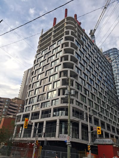 This is a low-angle, vertical shot of a modern, multi-story building under construction, likely in an urban setting in Ottawa, Canada. The building's facade is characterized by a grid of dark windows set within a repeating pattern of curved concrete balconies. A construction crane is visible in the upper left corner of the image, extending above the building. Power lines and utility poles are strung across the sky, partially obscuring the building and the sky. Traffic signals are also visible, including a yellow signal with three lights. One sign beneath it reads "BICYCLES EXCEPTED." Another sign on a pole has a circular symbol with a diagonal line through it, indicating "no" for something, and "RIM CONSTR" is visible on a sign on the building. The sky is cloudy, suggesting it could be overcast or a partly cloudy day. The presence of construction materials and equipment, along with the incomplete state of the building, indicates that it is an active construction site. The overall mood is one of urban development and ongoing activity.
