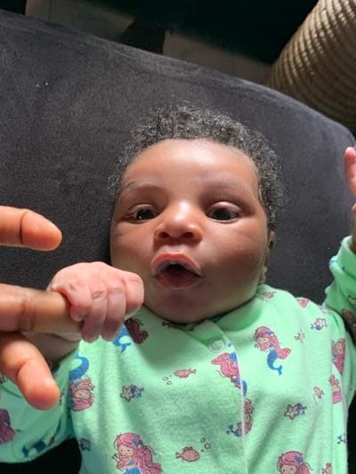 A close-up, slightly tilted image captures an infant lying on a dark grey or black textured surface, likely a blanket or furniture. The baby has dark, curly hair and dark skin, with wide, dark eyes looking directly at the viewer. The infant's mouth is slightly open, hinting at an expression of curiosity or an attempted sound.

The baby is dressed in a light green pajama or romper covered with whimsical patterns of mermaids, starfish, and small fish, primarily in pink, blue, and brown tones. The garment has visible snap buttons along the side.

In the foreground, the baby's small hand is firmly gripping an adult's index finger, showcasing a tender moment of connection and the infant's natural grasping reflex. Only a portion of the adult's hand is visible. The background, particularly in the upper left, features a blurred, textured object, possibly a woven basket or furniture piece, in a warm, earthy tone. The setting appears to be indoors, under ambient lighting. The scene conveys a sense of warmth and interaction between the infant and caregiver. No visible text is present. This image was taken in Ibasa, Nigeria.