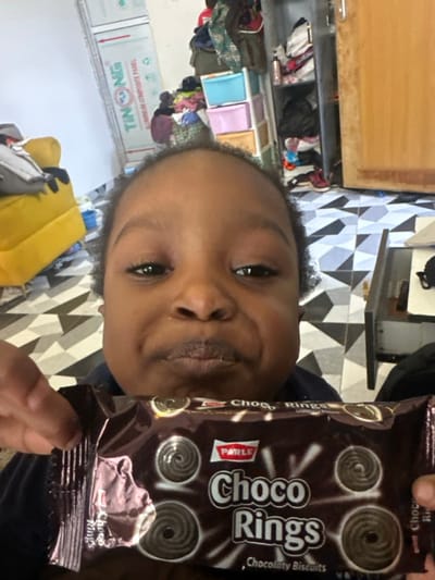 A young individual with dark complexion and short, dark hair, wearing a dark garment, is depicted in a medium shot, looking directly towards the camera with a slight pout. They are holding a brown, foil-wrapped package of "Parle Choco Rings" Chocolaty Biscuits in both hands, positioned below their chin. The background reveals an indoor residential setting in Ilishan, Nigeria. The floor features black and white geometric patterned tiles. To the left, a yellow upholstered armchair is partially visible. Further back and to the right, a multi-tiered plastic drawer unit, containing various items, stands next to a wooden door. A white wall section to the left displays a red and white sign for "TINONG ALUMINIUM COMPOSITE PANEL." The environment appears lived-in.