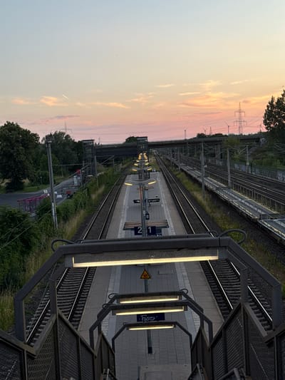 An outdoor, high-angle, full shot shows a train station with multiple tracks and platforms at dusk. The sky displays hues of pink and orange from the setting sun, with wispy clouds and a contrail visible. On the left side of the frame, a pink bus shelter with metal supports stands near lush green trees. The train tracks run parallel towards the horizon, bordered by platforms equipped with lighting fixtures and benches. Signage indicating platform numbers, "1" and "2," is visible on the platforms. To the right, a chain-link fence separates the tracks from overgrown vegetation and a graffiti-covered wall. In the background, a bridge crosses over the tracks, with power lines and wind turbines visible in the distance. The scene is deserted and lacks any discernible human presence or activity. The location is identified as the train station in Wustermark, Germany.