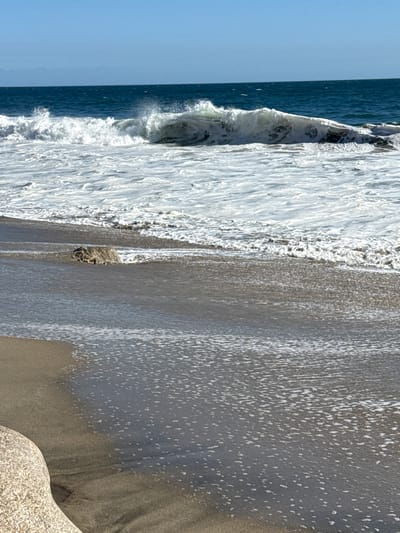 The image depicts a segment of the Pacific Ocean coastline in Malibu, California, United States of America. A powerful wave is captured mid-break, its crest white with foam as it crashes onto the shore. The water exhibits a deep blue hue, contrasting with the foamy white of the breaking wave and the lighter, agitated water closer to the beach. The foreground shows a sandy beach with shallow water lapping at the shore, creating ripples and small patches of foam on the wet sand. A large, light-colored rock is visible in the lower left portion of the image, partially submerged by the receding tide. The sky above is a clear, bright blue. The scene is devoid of any people or man-made structures, emphasizing the natural beauty of the coastal environment. The lighting suggests a sunny day, with the sun casting a gentle illumination on the water and sand.