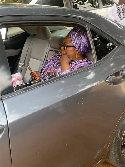 A woman wearing a purple patterned headwrap, glasses, and a matching purple patterned outfit, adorned with large gold earrings, is seated in the back of a grey sedan. She holds a smartphone in her right hand, angled towards her face as if interacting with it. Her left hand rests on her chin, and her gaze is directed downwards. To her left, a white plastic bag with pink accents sits on the car's seat. The interior of the car is visible, including grey upholstery and a seatbelt. Outside the car, greenery and what appears to be another vehicle are reflected in the windows. The car's exterior exhibits several scratches and scuff marks, particularly on the rear quarter panel, indicative of wear and tear. The scene is captured from a slightly elevated, exterior perspective, focusing on the woman and the immediate interior of the vehicle. This scene was photographed in the city of Riyom, Nigeria.