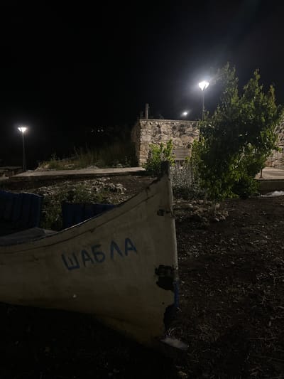 This is a nighttime photograph taken in Tyulenovo, Bulgaria, featuring a partially visible boat in the foreground. The boat is white with blue markings and has the Cyrillic word "ШАБЛА" painted on its side. The scene is dimly lit by streetlights, one of which is visible on the left and another further in the background. Behind the boat, a stone wall structure is visible, partially obscured by foliage and a tree. The ground is covered in dark soil and rocks. The overall atmosphere is quiet and somewhat desolate, characteristic of a remote coastal area at night.