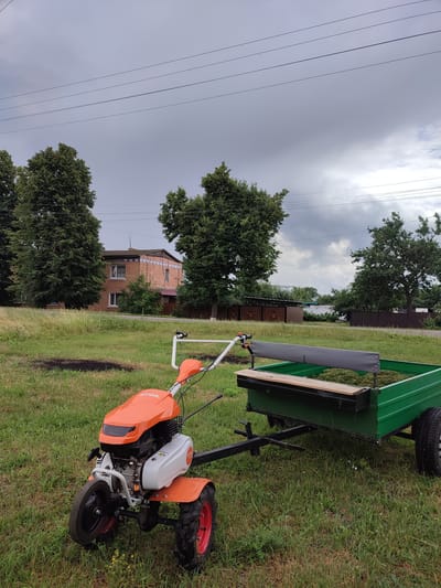 An orange and white Stihl tiller is hitched to a green metal trailer loaded with cut grass. The tiller is in the foreground, to the left, with its wheels on the grass. The trailer is to the right of the tiller, also on the grass, and appears to be a utility trailer with a wooden bench seat and a metal frame. Behind the trailer, there is a dark brown wooden fence and a white wall with a metal roof. Further in the background, there is a two-story brick building with a tiled roof and several large green trees. The sky above is cloudy and overcast, with power lines visible crisscrossing the upper portion of the image. The scene appears to be set in a rural or semi-rural area, possibly a backyard or a field, in the village of Voskobiinyky, Ukraine.