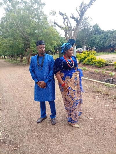 A man and a woman, dressed in traditional blue attire with gold accents, stand side-by-side on a dirt path in Tambes Mile Four, Nigeria. The man wears a long-sleeved tunic and trousers, a black patterned cap, and multiple strands of black beads around his neck. His hands are clasped in front of him. The woman is adorned in a velvet top with puffy sleeves and a long skirt featuring intricate gold embroidery. She wears a blue headwrap, a coral beaded necklace, and matching bracelets. Her left hand rests on her hip. Behind them, trees with sparse foliage and some greenery line the path. The ground is covered in small stones and dry leaves. The overall impression is of a formal occasion in an outdoor, possibly rural, setting.