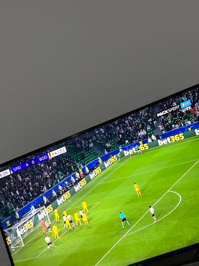A photograph taken in Sredkovets, Bulgaria, captures a television screen displaying a live football match, with a plain light gray wall or ceiling visible in the upper left portion of the frame. The television screen is angled, showing a brightly lit green football pitch with white markings. In the top left corner of the screen, a scoreboard reads "6:39 SPO 4 0 BOD (4 3)". The top right corner displays the channel logo "MAXSPORT 3 HD LIVE". A dense crowd of spectators occupies green stadium seats in the background. Along the touchline, electronic advertising boards repeatedly feature the "bet365" logo. On the field, multiple football players are visible: a group of players in yellow jerseys, including a goalkeeper, are gathered near a white goal, forming a defensive wall. Other players in black and white jerseys are positioned nearby. A player in a black and white jersey stands further from the goal, while a referee in a light blue jersey observes the scene.