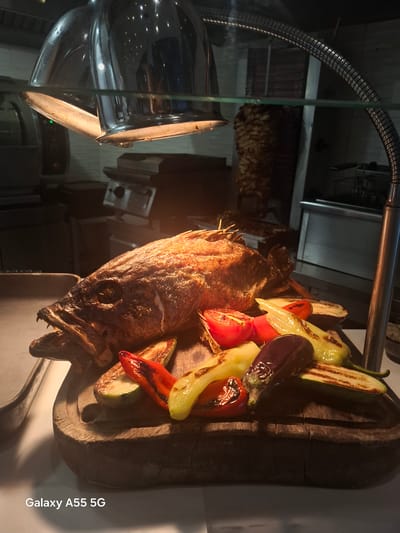 The image displays a beautifully arranged food presentation, likely within a restaurant or buffet setting in Gouves Municipal Unit, Greece. The primary subjects are a whole grilled fish and a colorful assortment of grilled vegetables, presented on a rustic wooden cutting board.

The fish, appearing to be a sea bream or similar species, is golden-brown with crispy skin, suggesting it is freshly cooked. Its head is visible on the left, facing forward, with an open mouth and visible teeth. Wooden skewers are visible, possibly holding the fish in place. To the right of the fish, on the same board, are various grilled vegetables, including vibrant red bell peppers, pale yellow peppers, dark purple eggplant slices, and green zucchini halves, all bearing distinct char marks from grilling.

A prominent metal heat lamp, positioned above and to the left, casts a warm, focused light on the food, indicating it is being kept warm for serving. Reflections from the lamp are visible on surrounding surfaces, including a glass or clear plastic barrier in the foreground. The background is dimly lit and slightly out of focus, revealing kitchen-like elements such as metallic surfaces and indistinct shapes, reinforcing the commercial food service context.

No people are visible in the scene. The artificial lighting suggests an indoor setting or an evening presentation. The text "Galaxy A55 5G" is visible vertically along the right side of the image, identifying the device used to capture the photograph. The overall impression is of fresh, appealing Mediterranean cuisine.