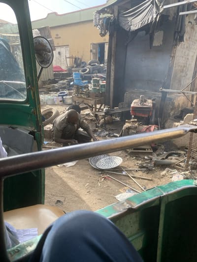 A man squats in a cluttered outdoor space, likely a shop or yard filled with mechanical parts and debris. He appears to be working, possibly on repairing something. The view is taken from inside a vehicle, possibly a tuk-tuk, with a green frame and a side mirror visible. The scene feels busy and slightly chaotic.