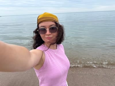 A woman stands on a beach in Derbent, Russia, posing for a selfie. She is wearing a pink knitted top, a yellow and brown bandana, and sunglasses. The sea is visible behind her, with a calm surface under a cloudy sky. The beach is made of sand and small pebbles.