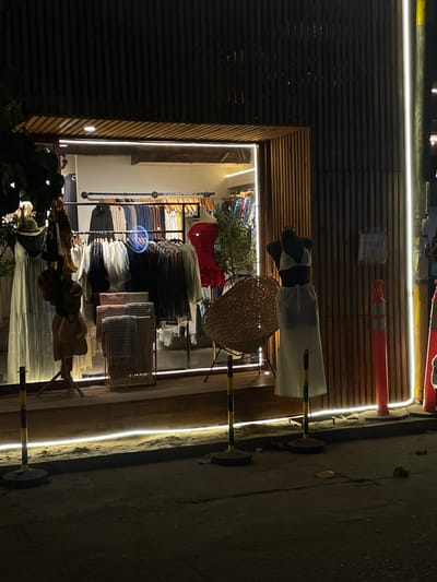 A shop in Brisas de Zicatela, Mexico, displays clothing and accessories. A mannequin dressed in a white dress and black and white top is visible, along with a straw hat. Racks of clothing and hats are on display within the shop's interior. The shopfront is lit with LED strip lights. The exterior is characterized by horizontal wooden slats. Part of the street is visible in the foreground.