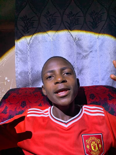 A young Black male is seated in a red chair with a patterned design. He is wearing a red Manchester United jersey with white stripes on the shoulders and the club's crest on the chest. The background consists of a dark patterned curtain and a lighter patterned curtain, with a band of light and shadow across the top. The lighting creates a dramatic effect with strong shadows and highlights. The overall scene appears to be an indoor setting, possibly a home, in Barakin Sabawa, Nigeria.
