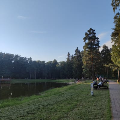 The image shows a serene park scene in Il'inskiy, Russia, with a calm lake reflecting the surrounding tall pine trees and a clear blue sky. A grassy embankment slopes down to the water's edge, with recently cut grass clippings visible. On the right side of the image, a paved walkway runs alongside the grassy area, bordered by trees. Several people are visible in the park, some sitting on a bench near the walkway, while others are seen in the distance near the lake. Bicycles are parked near the bench, suggesting recreational activity. A wooden pier extends into the lake on the left side of the image. The overall atmosphere is peaceful and suggests a pleasant day in a natural setting within the city.