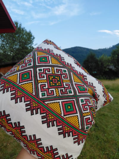 The image shows a close-up of a white pillow with traditional Ukrainian embroidery, held up outdoors. The embroidery features geometric patterns in red, yellow, green, and black. In the background, out of focus, are trees, a roof of a wooden building, green grass, and rolling hills under a cloudy blue sky. The image appears to be taken in a rural or mountainous area, consistent with parts of Ukraine like Chernyk. There are no people visible in the image, and the focus is entirely on the embroidered pillow.