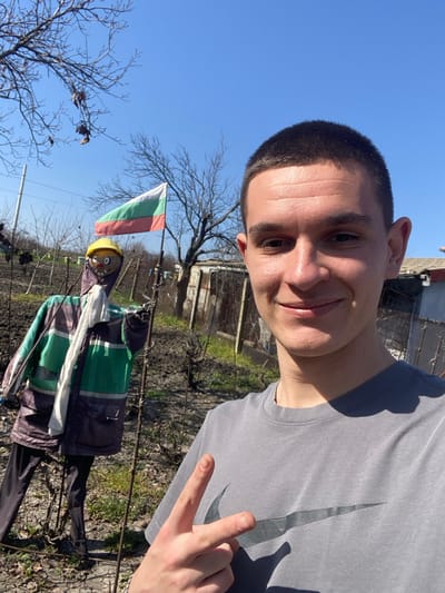 A young male, with short dark hair and a grey Nike t-shirt, is shown in a frontal selfie. He is smiling and displaying a two-finger peace gesture with his right hand. The scene is an outdoor setting, identified as a garden or cultivated area within the city of Merichleri, Bulgaria. Behind the male, a scarecrow is positioned to the left, dressed in a yellow hard hat, a multi-paneled jacket featuring green, grey, and dark blue/purple sections with reflective elements, a white scarf, and dark trousers. A Bulgarian flag, horizontally striped in white, green, and red, is visible on a pole further back, to the right of the scarecrow. The environment consists of disturbed soil with sparse green growth, several bare deciduous trees, and rudimentary structures with fencing, all under a clear blue sky.
